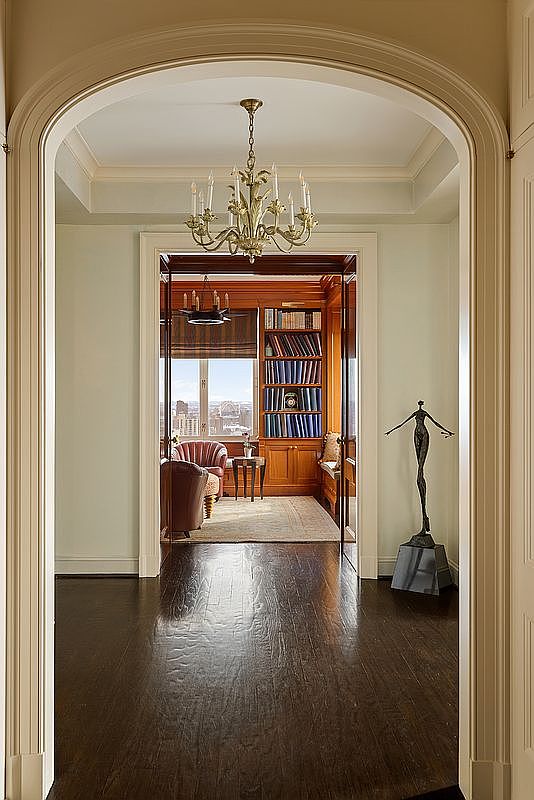 This elegant hallway features a grand arched entryway leading into a sophisticated wood-paneled library. The foreground showcases dark hardwood floors and a striking abstract sculpture, while a classic chandelier hangs from the ceiling, creating a refined and luxurious atmosphere. The perspective draws the eye through the transition space into the warm, inviting study beyond, which offers a scenic city view.