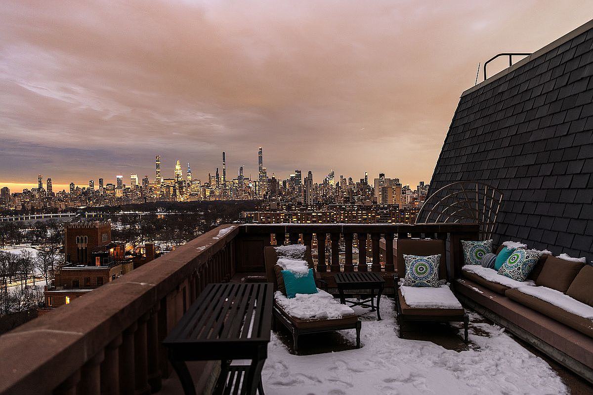This stunning rooftop terrace offers a breathtaking, panoramic view of the New York City skyline at dusk, framed by a dusting of fresh snow. The space is furnished with comfortable lounge seating and a dining table, creating an inviting outdoor retreat despite the winter chill. The cinematic perspective captures the contrast between the cozy, snow-covered furniture and the vast, glowing urban horizon.