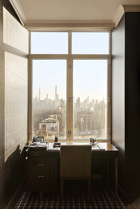 This sophisticated home office features a dark, polished desk positioned directly in front of a large, multi-pane window that frames a stunning, expansive city skyline view. The room is framed by dark, textured wall paneling and a patterned carpet, creating a moody and professional atmosphere. The perspective is centered and symmetrical, emphasizing the desk as a focal point against the bright, urban backdrop.