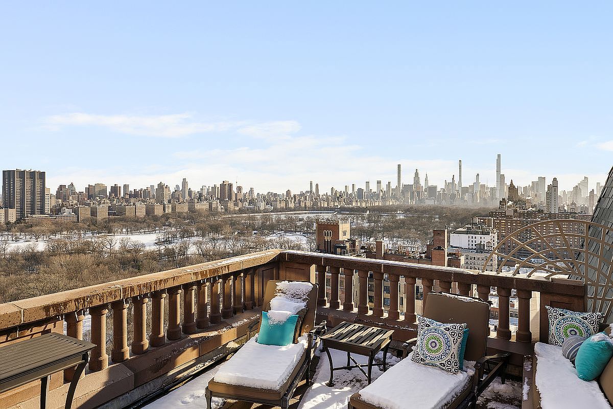 This high-rise balcony offers a breathtaking, panoramic view of Central Park and the iconic New York City skyline under a clear winter sky. The outdoor space is furnished with lounge chairs topped with light snow, featuring decorative patterned pillows that add a touch of color to the wintry setting. The stone balustrade and elegant architectural details frame the expansive urban vista, creating a sophisticated and serene atmosphere.