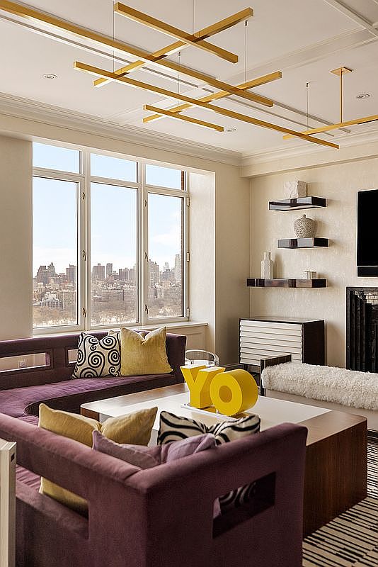This sophisticated living room features a plush, deep purple sectional sofa accented with patterned and solid gold throw pillows, centered around a modern coffee table displaying bold yellow 'YO' decor. A striking, geometric gold chandelier hangs from the ceiling, while large windows provide a stunning, expansive view of the city skyline. The space is further enhanced by minimalist floating shelves and a sleek, contemporary fireplace, creating an atmosphere of urban luxury and refined comfort.