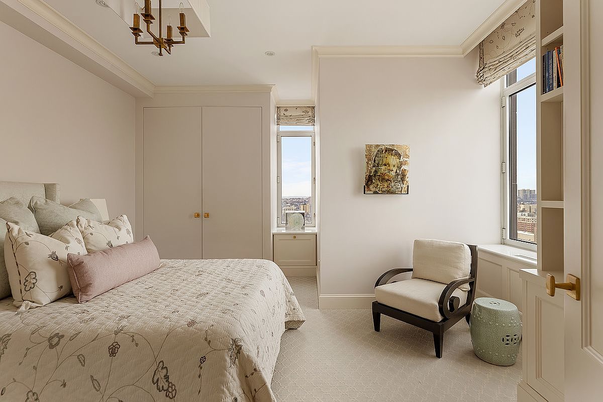 This elegant and serene guest bedroom features a neutral color palette, a plush bed with floral-patterned bedding, and a cozy seating area by the window. The room is accented by a unique piece of wall art, built-in shelving, and a classic chandelier, creating a sophisticated and inviting atmosphere. The perspective is a wide-angle shot that captures the entire room, highlighting the blend of comfort and refined design.