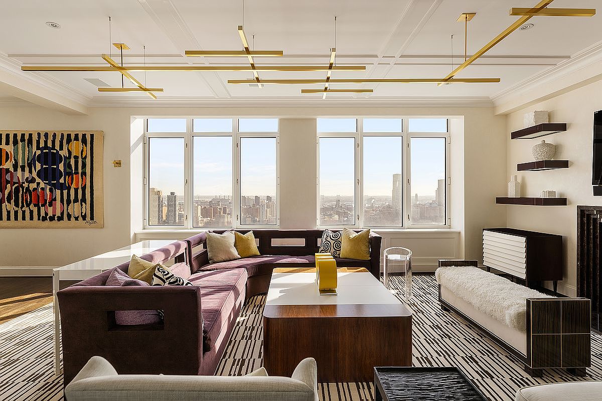This sophisticated living room features a large, plush purple sectional sofa arranged around a modern, two-toned coffee table, set against a backdrop of expansive windows offering a panoramic city view. The room is accented by a striking geometric gold light fixture suspended from the ceiling, a patterned area rug, and minimalist shelving units. The overall aesthetic is luxurious and contemporary, blending bold textures with a clean, high-end design.