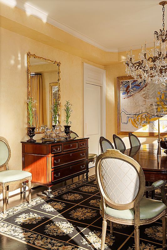 This elegant dining room features a rich, dark wood buffet topped with a decorative silver service and a large, ornate gold-framed mirror. A sophisticated black and gold patterned rug anchors the space, while a crystal chandelier hangs above a dining table, casting a warm glow over the light-colored walls. The room is furnished with classic, upholstered chairs, creating a formal and luxurious atmosphere perfect for refined entertaining.