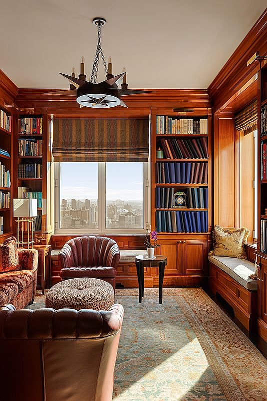 This sophisticated home library or study features rich, floor-to-ceiling wood paneling and built-in bookshelves filled with volumes. The room is anchored by a plush, dark leather armchair and a matching ottoman, complemented by a patterned area rug and a unique star-shaped chandelier. A large window provides a stunning, elevated city skyline view, creating a warm, scholarly atmosphere perfect for quiet reflection.
