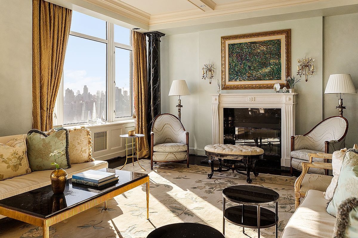 This elegant, sun-drenched living room features a sophisticated blend of classic and contemporary design, highlighted by a grand fireplace and a large window offering a stunning city skyline view. The space is furnished with plush, patterned armchairs, a sleek coffee table, and a neutral-toned area rug, all anchored by a striking black marble column. The overall atmosphere is one of refined luxury and comfort, perfect for high-end urban living.