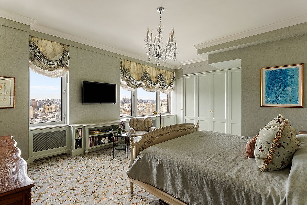 This elegant primary bedroom features a classic, sophisticated design with soft sage-green walls, a patterned carpet, and a large bed adorned with decorative pillows. The room is bathed in natural light from two large windows that offer expansive city views, complemented by a crystal chandelier and built-in storage cabinetry. The overall atmosphere is serene and refined, blending traditional comfort with a high-rise urban perspective.