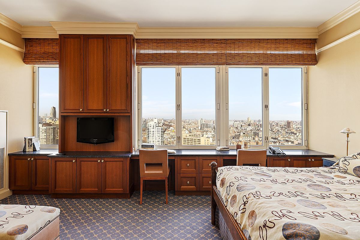 This bedroom features a built-in wooden desk and cabinetry unit that spans the length of the window wall, offering a functional workspace with an expansive city view. The room is furnished with a bed featuring a patterned comforter and is carpeted with a blue, patterned rug. The overall aesthetic is traditional and professional, emphasizing the high-rise urban location.