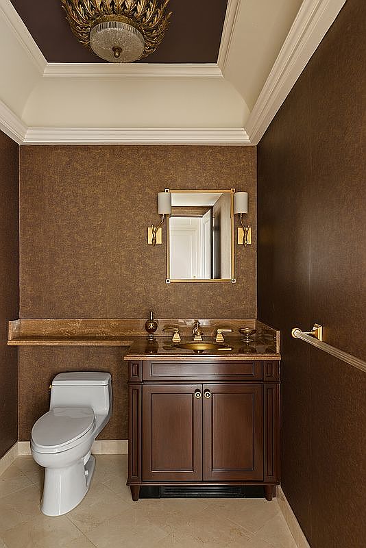This elegant powder room features rich, textured brown wallpaper, a dark wood vanity with a gold-toned sink, and a matching gold-framed mirror. The space is accented by a sophisticated, ornate ceiling light fixture and a white toilet, creating a luxurious and moody atmosphere. The perspective is a straight-on, eye-level shot that highlights the high-end finishes and compact, functional layout.