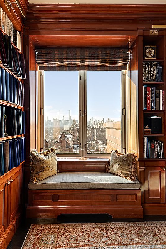 This sophisticated study features rich, dark wood built-in bookshelves flanking a cozy window seat that offers a stunning, elevated view of the city skyline. The space is accented by elegant patterned throw pillows and a classic area rug, creating a warm and scholarly atmosphere. The perspective is centered on the window nook, emphasizing the luxurious craftsmanship and the impressive urban vista.