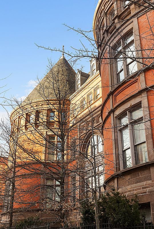 This image captures a striking side view of a historic, red-brick building featuring a prominent conical turret and intricate architectural detailing. The perspective is angled upward from the street level, emphasizing the building's height and the texture of the masonry against a clear blue sky. Bare tree branches frame the structure, adding a sense of seasonal character and depth to the composition.