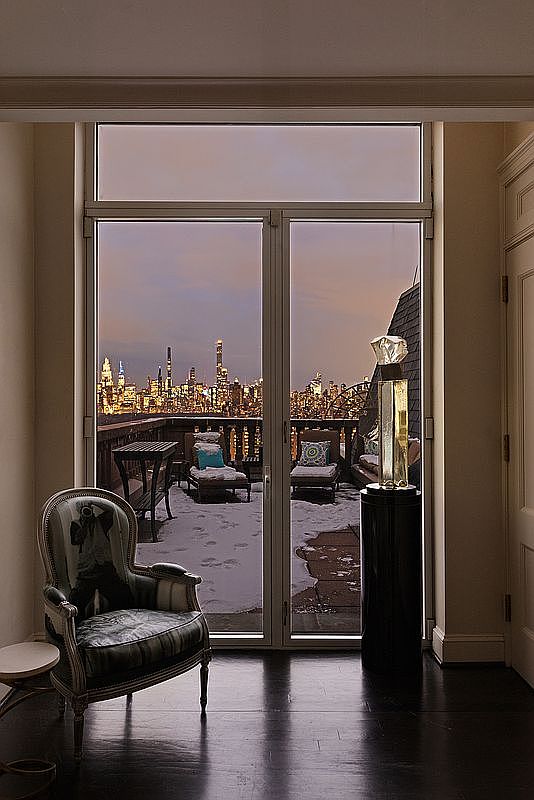 This sophisticated interior space features a classic armchair positioned near a large glass door that opens onto a snowy terrace with a breathtaking panoramic view of the city skyline at dusk. The room is elegantly furnished with a dark, polished floor and a sculptural art piece on a pedestal, creating a luxurious and serene atmosphere. The cinematic perspective frames the urban landscape through the doorway, emphasizing the contrast between the warm, refined interior and the cold, vibrant city lights outside.