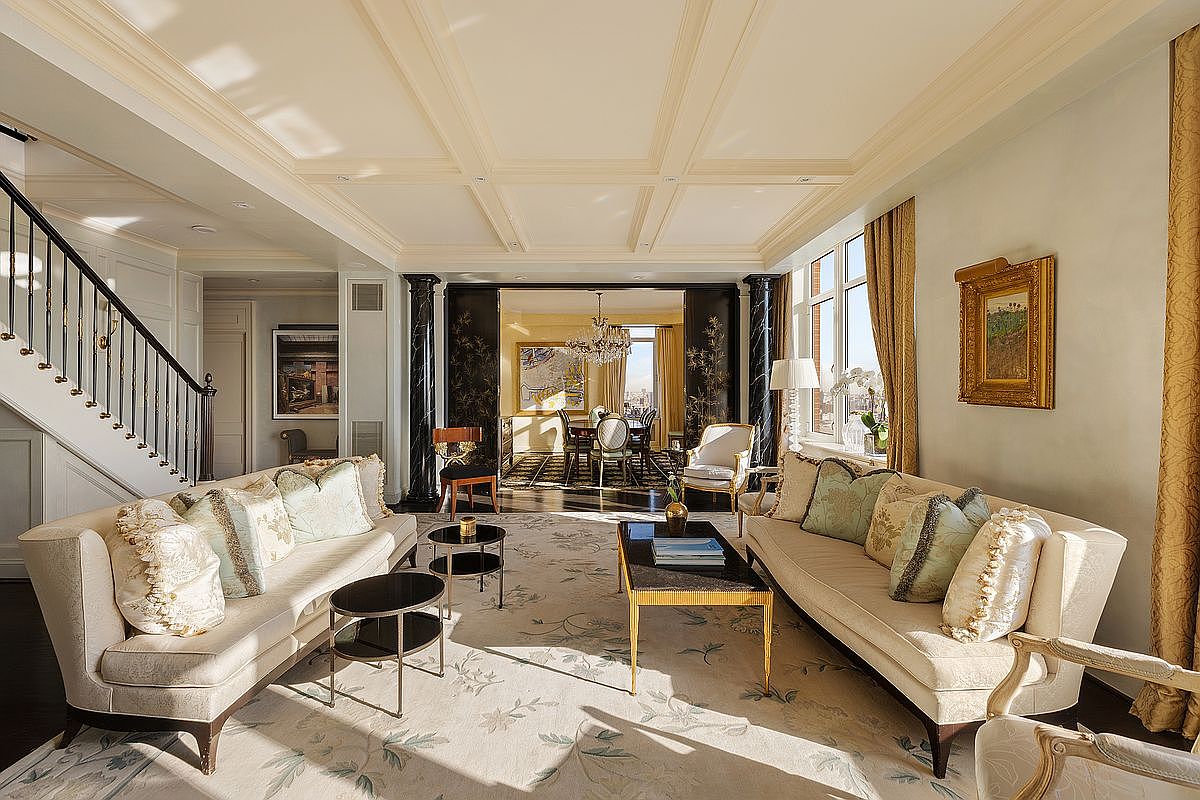 This elegant, formal living room features a sophisticated neutral color palette with high-end furnishings, including two plush sofas and a central coffee table. The space is defined by intricate coffered ceilings, dark wood flooring, and a grand staircase, leading into an adjacent dining area framed by striking black columns. Large windows draped in gold curtains allow natural light to fill the room, creating a luxurious and inviting atmosphere.