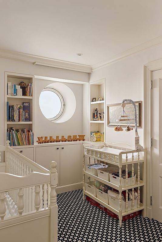 This charming nursery features a classic white crib and a matching changing table, set against a backdrop of built-in shelving and cabinetry. A unique circular window serves as a focal point, while the bold, geometric black-and-white patterned carpet adds a modern touch to the traditional decor. The room is bright and well-organized, creating a cozy and inviting atmosphere for a child.