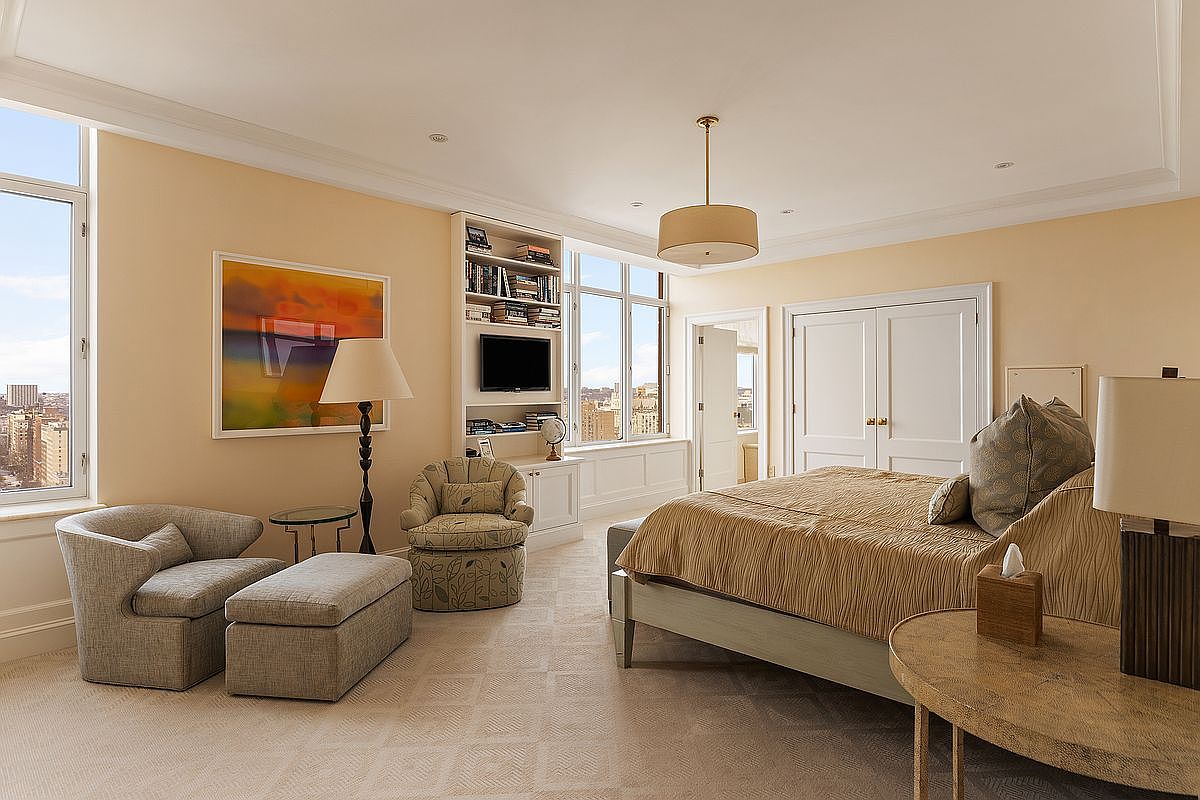This elegant primary bedroom features a neutral color palette, plush carpeting, and a large bed with textured bedding. The room is bathed in natural light from expansive windows that offer a stunning city view, complemented by a cozy seating area and built-in shelving. The overall atmosphere is sophisticated and serene, perfect for a high-end residential listing.