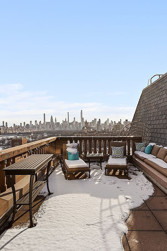 This expansive rooftop terrace offers a breathtaking, panoramic view of the New York City skyline, framed by a classic stone balustrade. The space is furnished with comfortable lounge seating and a sleek console table, all lightly dusted with a fresh layer of snow that adds a serene, wintery atmosphere. The perspective captures the contrast between the intimate, private outdoor living area and the vast, iconic urban landscape beyond.