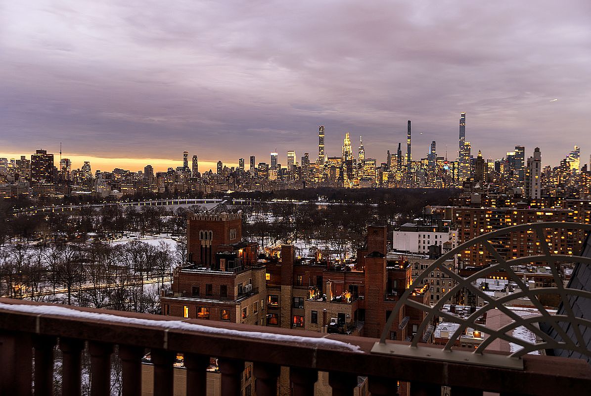 This image captures a breathtaking, elevated view from a balcony overlooking a snow-dusted Central Park at twilight. The foreground features a decorative metal railing and the architectural details of nearby brick buildings, while the background showcases the iconic, illuminated New York City skyline. The perspective is cinematic and expansive, emphasizing the luxury and prime location of the property.