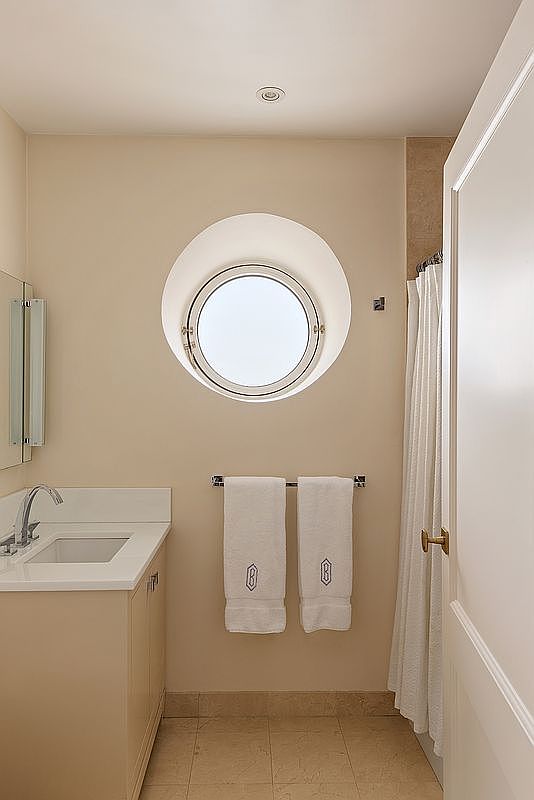 This clean and well-lit bathroom features a neutral color palette, a vanity with a white countertop, and a distinctive circular porthole-style window. A towel rack holds two white monogrammed towels, while a shower with a white curtain is positioned to the right. The space is simple and functional, offering a bright and airy atmosphere.