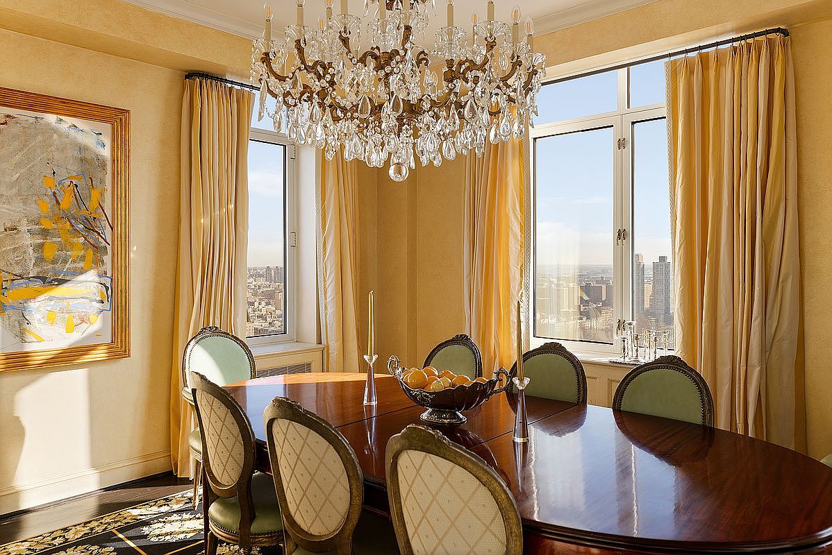 This elegant dining room features a polished mahogany oval table surrounded by classic upholstered chairs with light green accents. A grand crystal chandelier serves as the focal point above, while floor-to-ceiling yellow curtains frame expansive windows that offer a stunning, high-rise urban cityscape view. The room is further enhanced by warm, textured walls and a large, vibrant abstract painting, creating a sophisticated and luxurious atmosphere.