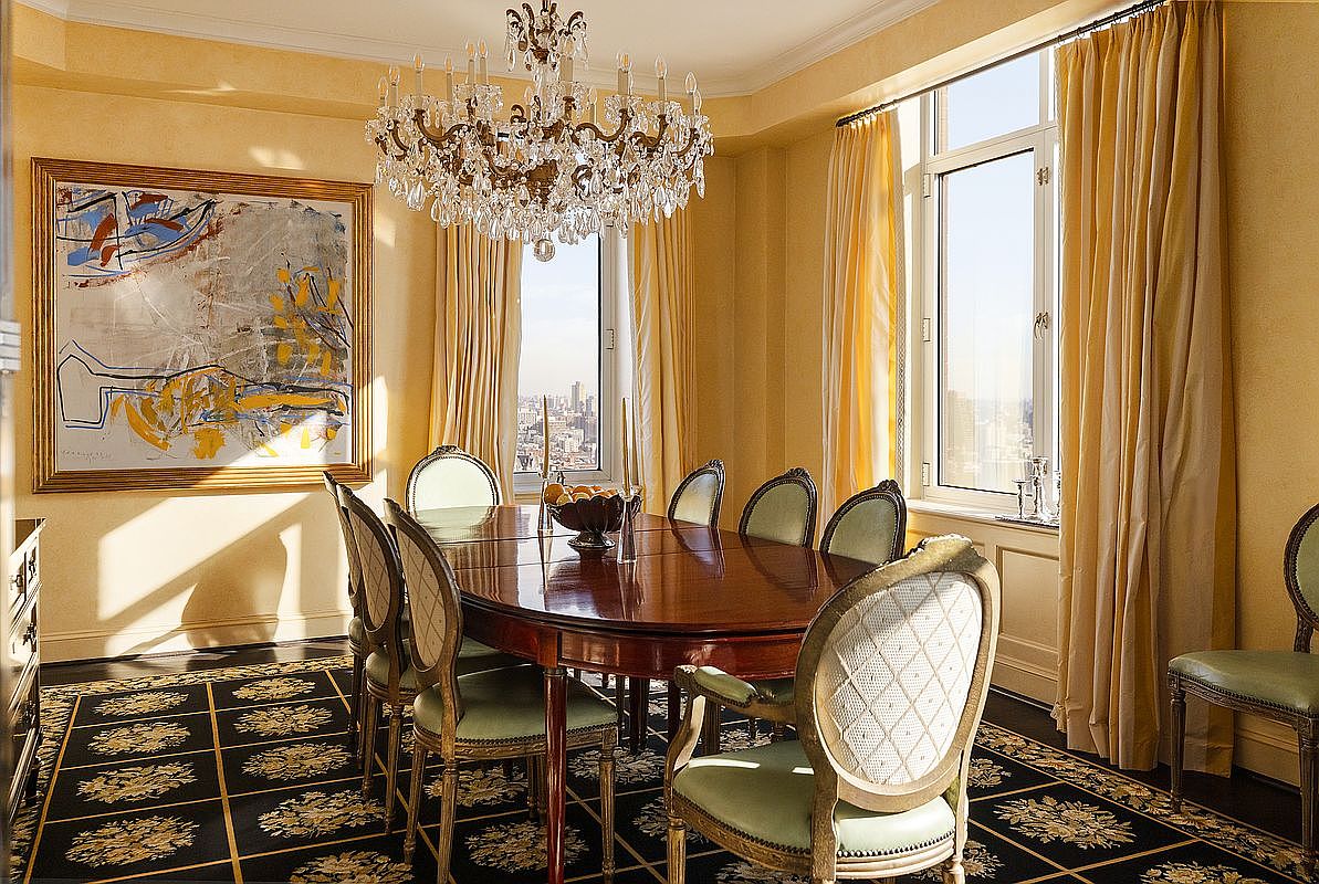 This elegant dining room features a polished mahogany table surrounded by upholstered chairs with light green fabric, set against a backdrop of warm yellow walls and floor-to-ceiling curtains. A grand crystal chandelier serves as the focal point above the table, while a large abstract painting adds a contemporary artistic touch to the space. The room is framed by expansive windows that offer a stunning, high-rise urban view, creating a sophisticated and luxurious atmosphere.