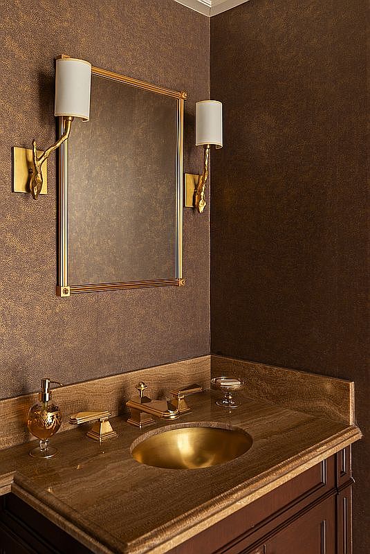 This elegant powder room features a rich, textured brown wallpaper that complements the warm, metallic tones of the brass sink and matching wall sconces. A framed mirror is centered between two ornate, branch-inspired light fixtures, while the vanity is topped with a polished stone countertop. The overall aesthetic is sophisticated and moody, creating a luxurious atmosphere.