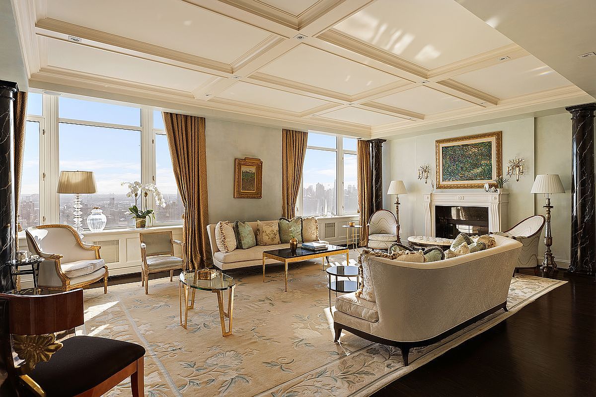 This elegant, high-end living room features a sophisticated neoclassical design with a coffered ceiling, dark hardwood floors, and expansive windows offering panoramic city views. The space is furnished with plush, cream-colored sofas, ornate accent chairs, and gold-trimmed coffee tables, all anchored by a large, intricate area rug. A grand fireplace with a framed painting above serves as the focal point, creating an atmosphere of refined luxury and timeless comfort.