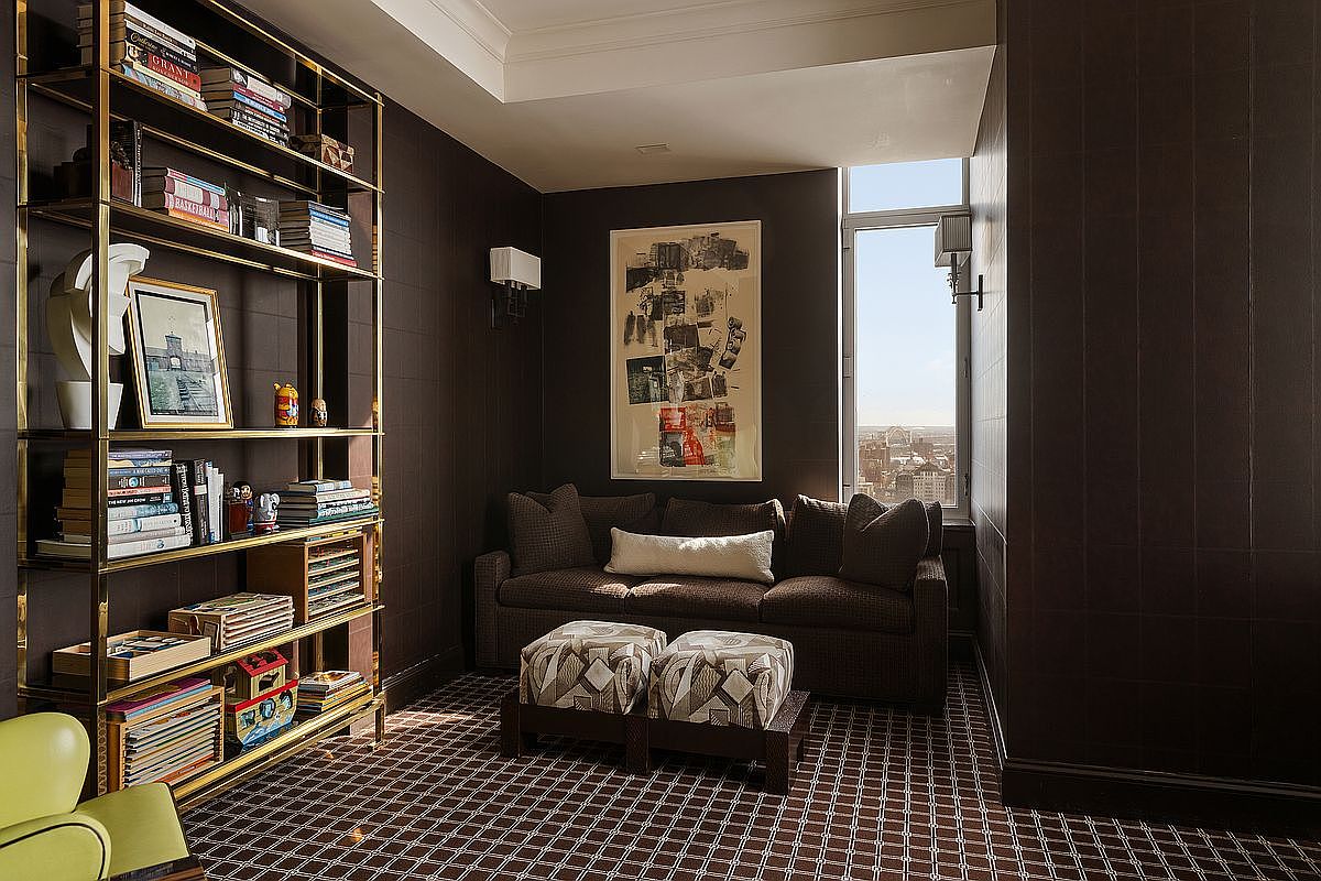 This sophisticated study features dark, textured walls and a rich, patterned carpet that creates an intimate and cozy atmosphere. A tall, gold-framed bookshelf filled with books and decorative items stands prominently on the left, while a plush sofa with patterned ottomans provides a comfortable seating area. A large window allows natural light to filter in, offering a view of the cityscape beyond.