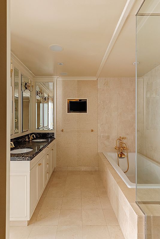This elegant primary bathroom features a double vanity with dark marble countertops, white cabinetry, and gold-toned fixtures. A deep soaking tub is positioned alongside a glass-enclosed shower, all set against neutral-toned stone tile walls and flooring. The space is well-lit with recessed lighting and includes a wall-mounted television, creating a luxurious and spa-like atmosphere.