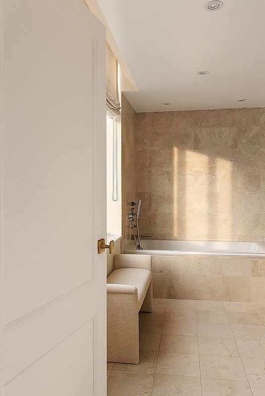This elegant bathroom features warm, neutral-toned stone tiling throughout the walls and floors, creating a serene and cohesive aesthetic. A built-in bench sits beside a deep soaking tub, which is bathed in soft, natural light streaming from a nearby window. The perspective is framed through a partially open white door, offering a welcoming and sophisticated glimpse into this tranquil retreat.