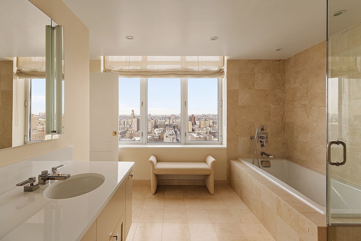 This elegant primary bathroom features a clean, contemporary design with light-toned stone tiling throughout. The space includes a sleek vanity with a white countertop, a deep soaking tub, and a central bench positioned in front of a large window that offers a stunning, expansive city view. The room is bathed in natural light, creating a serene and luxurious atmosphere.