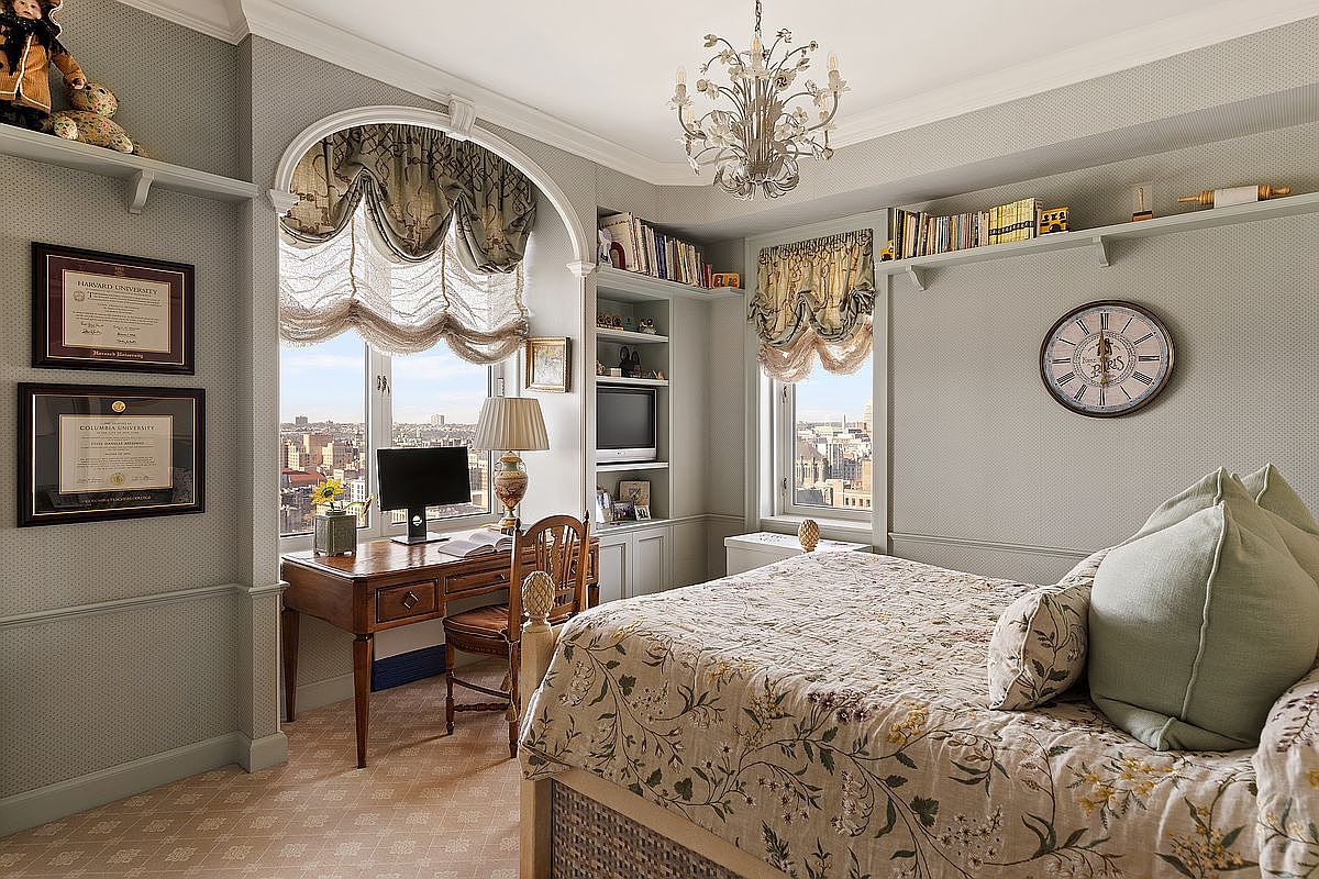 This elegant bedroom features a classic, traditional aesthetic with a floral-patterned bedspread, a sophisticated crystal chandelier, and custom built-in shelving. A dedicated workspace is positioned by the window, offering a stunning high-rise city view, while framed university diplomas on the wall add a scholarly touch. The room is bathed in natural light, creating a refined and cozy atmosphere suitable for a guest room or study.