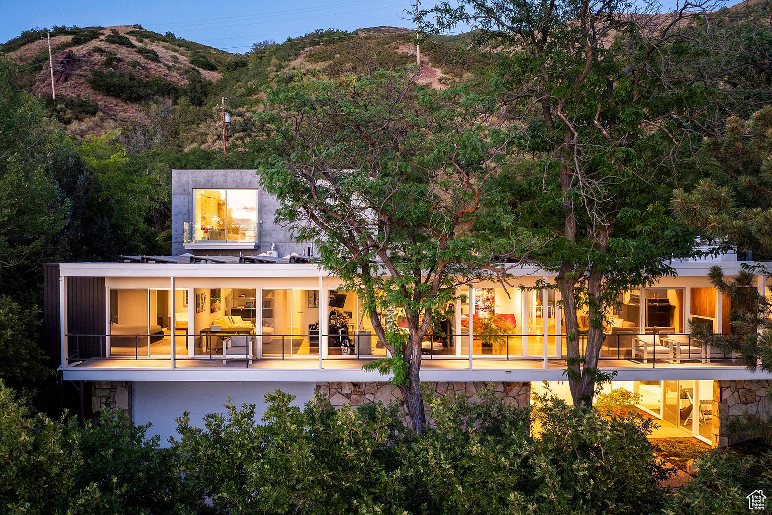 This is a front view of a modern, two-story house with a unique architectural design. The upper level features a wall of glass windows and a balcony, offering a bright and open living space. The lower level is constructed with stone, adding a touch of natural texture to the facade. The house is surrounded by lush greenery and trees, creating a sense of privacy and tranquility.