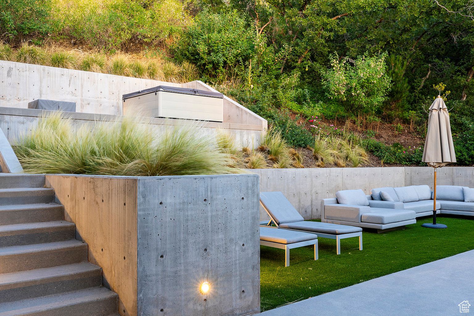 This image showcases a modern backyard design with tiered concrete retaining walls and lush greenery. A hot tub is nestled into the landscape, while a lounge area with sleek furniture and an umbrella offers a relaxing space. The artificial turf adds a touch of vibrancy and low maintenance appeal.