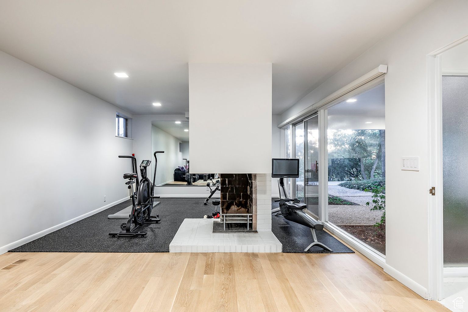 This is an interior shot of a home gym featuring a fireplace, exercise equipment, and a large sliding glass door that provides natural light and a view of the outdoors. The gym has a combination of hardwood and rubber flooring, with the exercise equipment placed on the rubber flooring. The perspective is from the entrance of the room, showcasing the layout and features of the space.