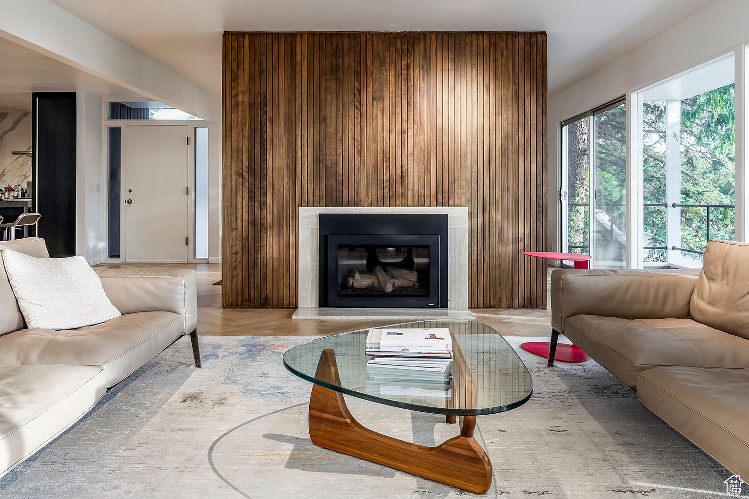 This living room features a striking wood-paneled wall with a modern fireplace, creating a warm and inviting focal point. Two beige leather sofas flank a glass-topped coffee table, set upon a patterned area rug. Large windows provide natural light, enhancing the room's spacious feel and contemporary design.