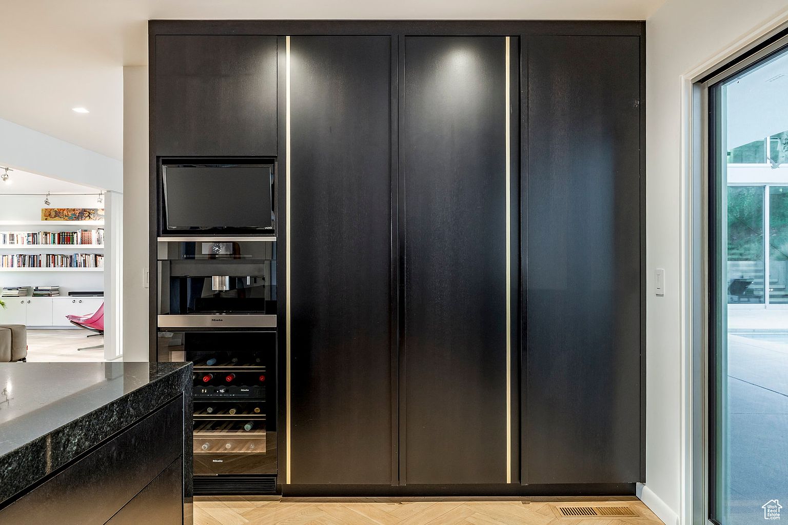 This image showcases a modern kitchen with sleek, dark cabinetry accented by vertical lighting strips. Integrated appliances, including a wine cooler, coffee maker, and television, are seamlessly incorporated into the cabinetry. The herringbone wood flooring adds a touch of warmth to the contemporary design.