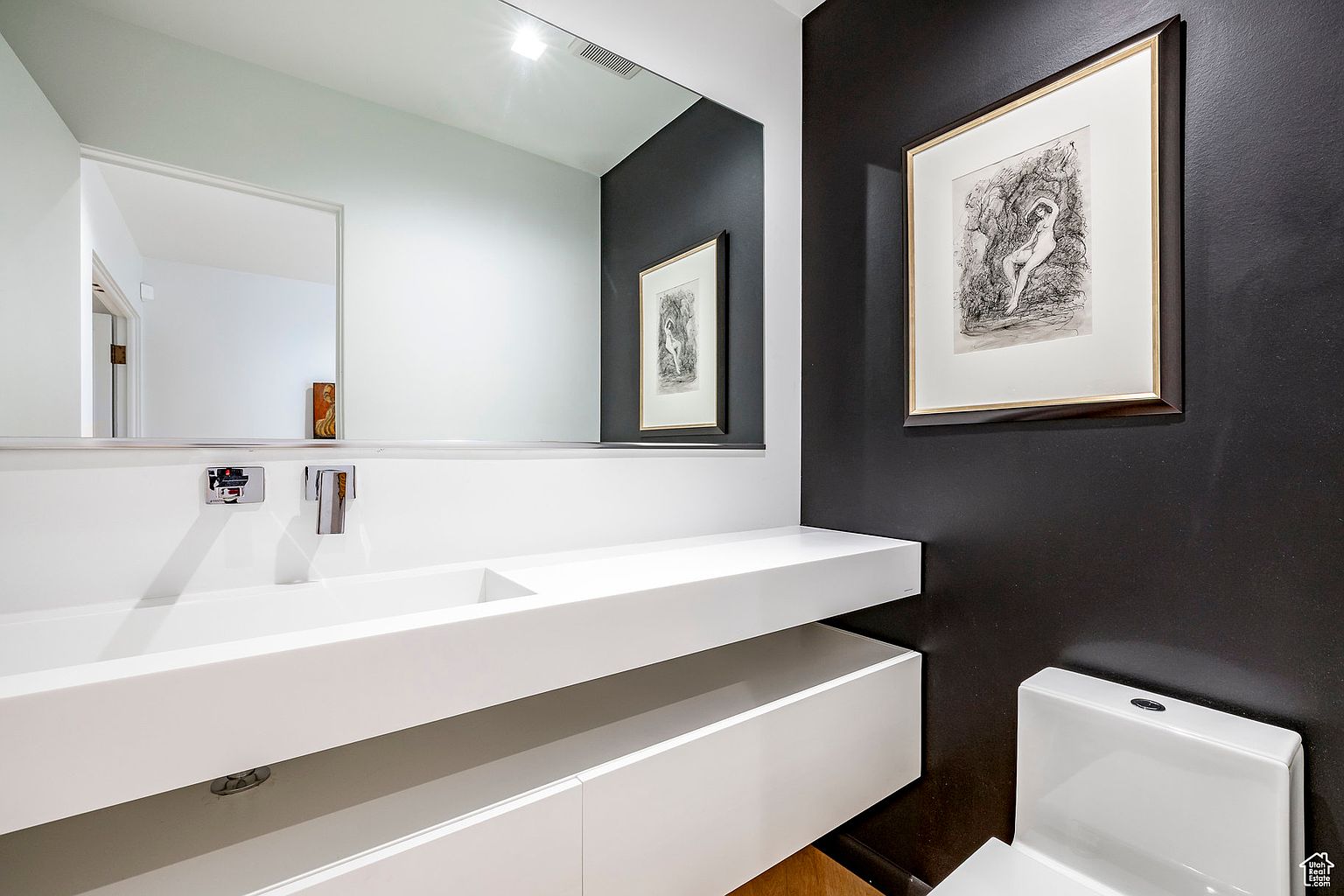 This is a modern guest bathroom featuring a sleek, floating white vanity with a rectangular sink and chrome fixtures. A large mirror reflects the vanity and a portion of the adjacent wall, which is painted a dark color and adorned with framed artwork. The overall design is minimalist and sophisticated, creating a clean and inviting space.