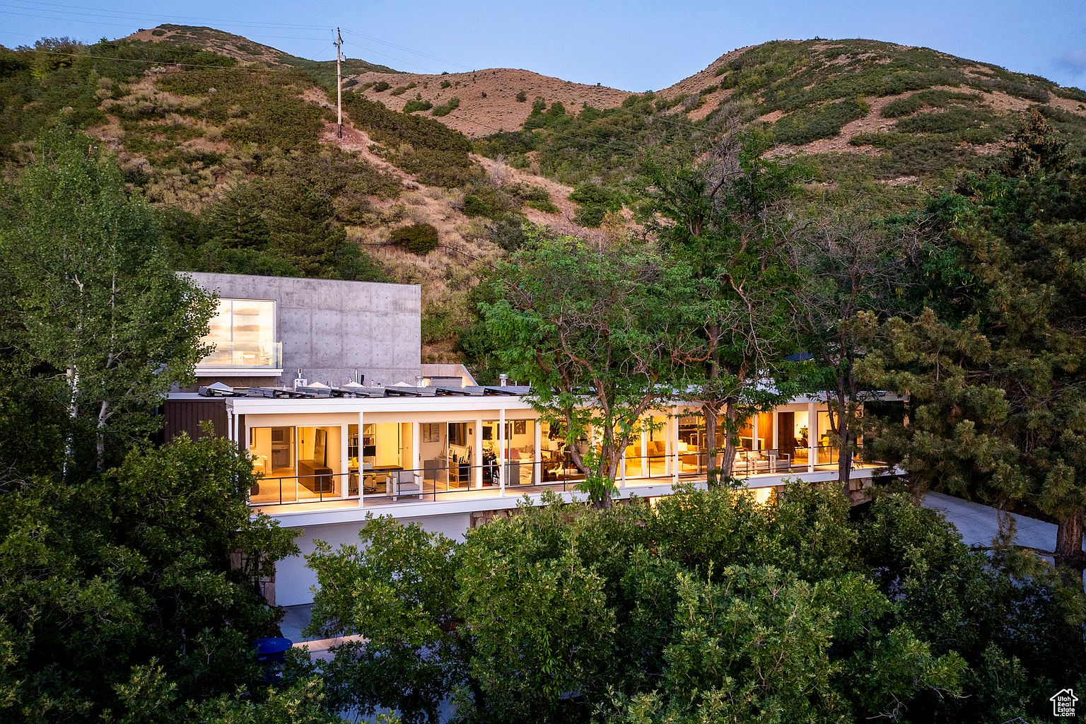 This is a rear view of a modern, multi-level home nestled into a hillside. The house features a long, glass-walled balcony or deck that spans the length of the structure, offering views of the surrounding landscape. The architecture incorporates concrete and wood elements, blending with the natural environment.