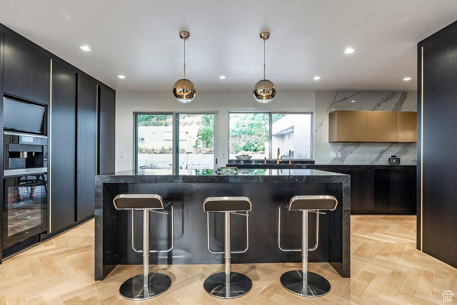 This is a modern kitchen featuring a large black island with seating for three. The kitchen includes black cabinetry with gold accents, stainless steel appliances, and pendant lighting. Large windows provide natural light and a view of the outdoors, while the herringbone wood flooring adds warmth to the space.