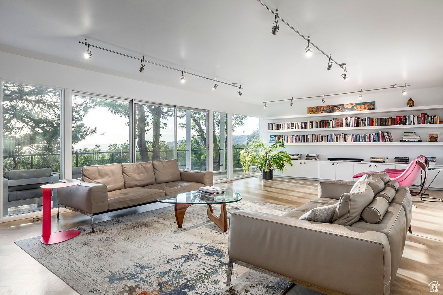 This is a bright and spacious living room featuring large windows that offer scenic views of the surrounding landscape. The room is furnished with two modern sofas, a glass-top coffee table, and a stylish area rug. A built-in bookshelf lines one wall, adding a touch of sophistication and functionality to the space.