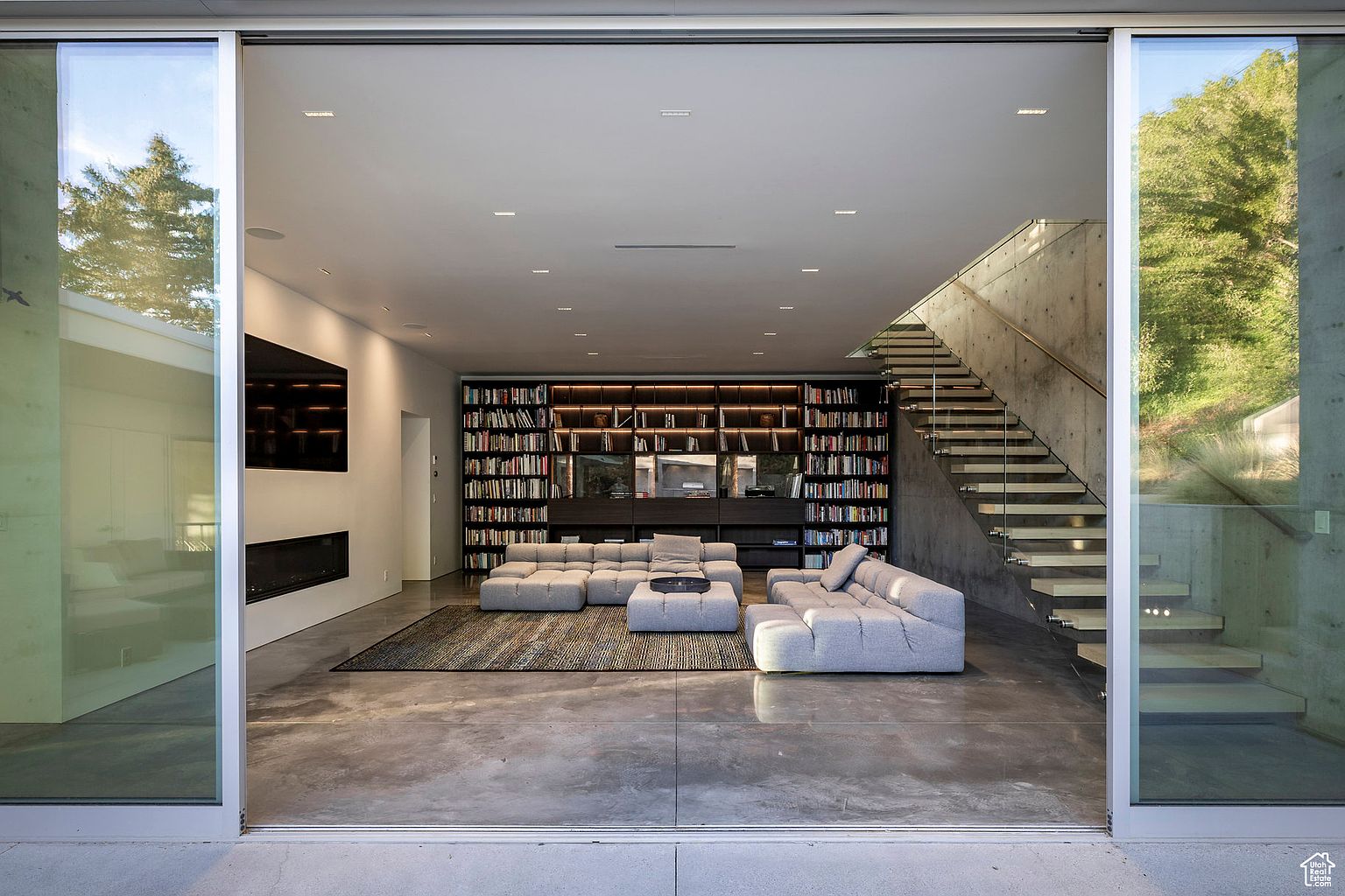 This is a spacious, modern living room featuring a large built-in bookshelf, a modular sofa, and a concrete floor. The room is well-lit with recessed lighting and natural light from the large sliding glass doors, which offer a view of the outdoors. A staircase with glass railings adds to the contemporary design.