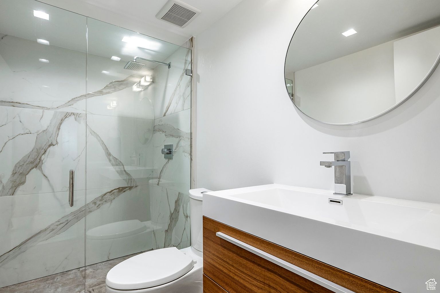 This is a modern bathroom featuring a glass-enclosed shower with marble-patterned tiles and a rainfall showerhead. The vanity has a sleek, white countertop with a wood-grain cabinet and a contemporary faucet. A round mirror hangs above the sink, and the overall aesthetic is clean and minimalist.