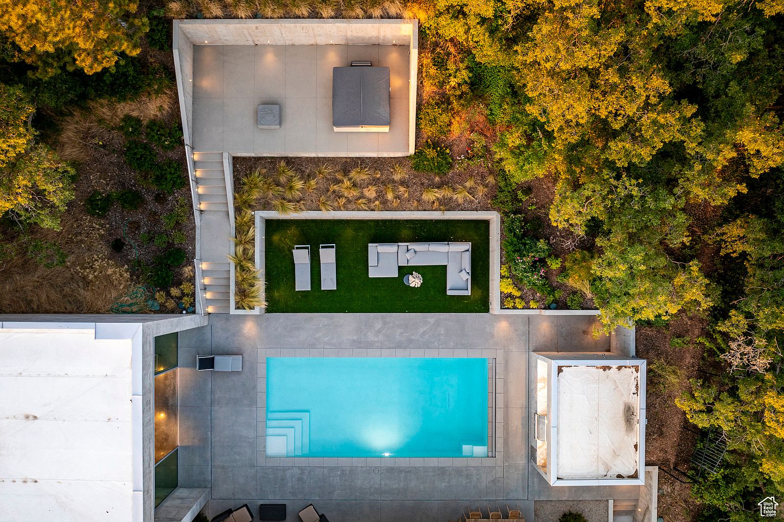 This aerial shot showcases a modern home's outdoor living space, featuring a rectangular swimming pool with a concrete surround, a lounge area with outdoor seating, and a separate patio with a hot tub. The landscaping includes manicured lawns, decorative grasses, and mature trees, creating a private and luxurious backyard retreat. The perspective is directly overhead, providing a comprehensive view of the property's layout and design.