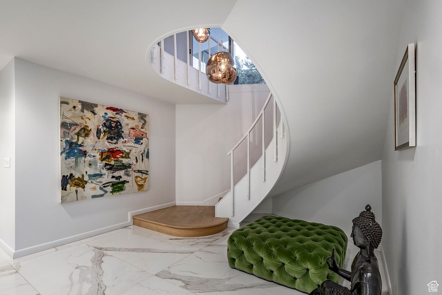 This interior shot showcases a modern hallway with a curved staircase. The space is bright and airy, featuring white walls, marble flooring, and a large abstract painting. A green tufted ottoman and a Buddha statue add a touch of elegance and character to the area.