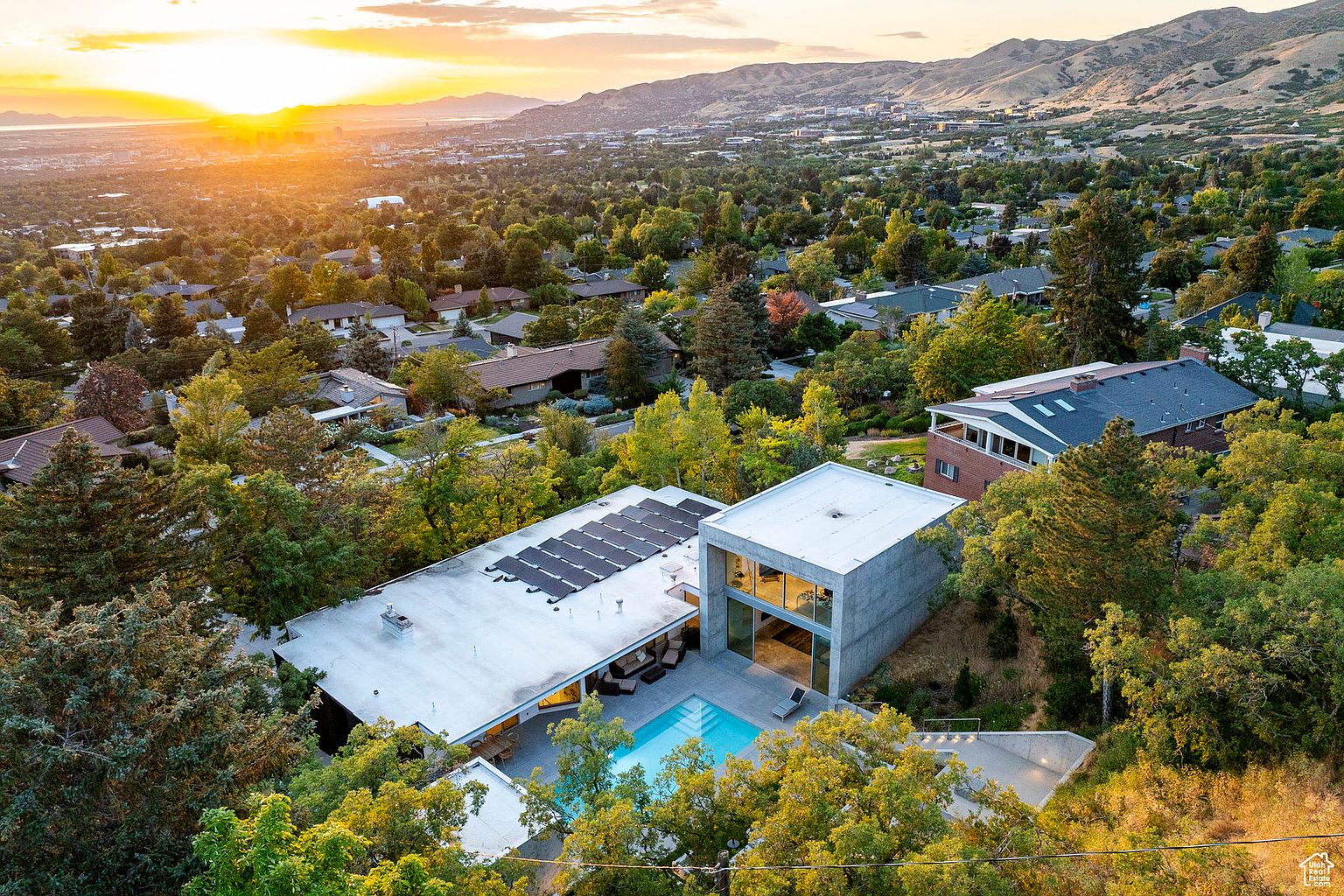 This aerial view showcases a modern home with a flat roof, solar panels, and a striking concrete cube structure. A swimming pool is nestled in the backyard, surrounded by lush greenery. The property offers a blend of contemporary design and natural beauty, with a stunning sunset backdrop over the city and mountains.