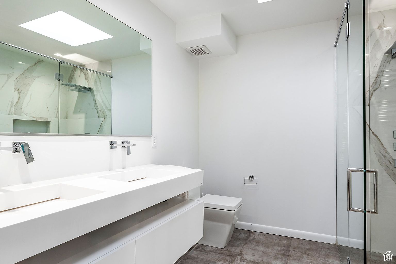 This is a modern primary bathroom featuring a double vanity with white countertops and cabinetry. A large mirror reflects the shower area, which is enclosed with glass and has marble-like tiling. The overall impression is clean, bright, and luxurious, with a minimalist aesthetic.