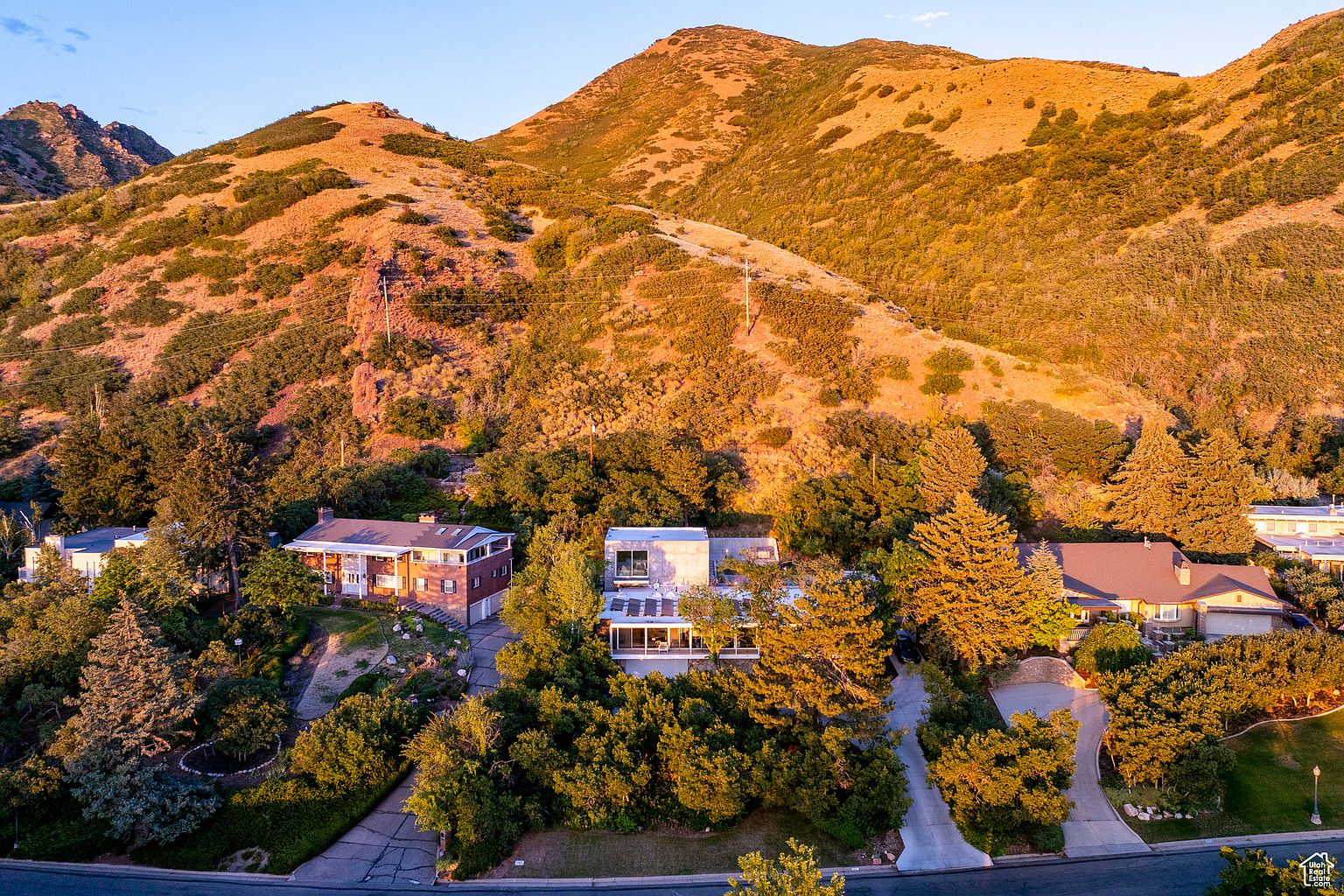 This aerial view showcases three distinct homes nestled amidst lush greenery and a backdrop of rolling hills. The homes feature varying architectural styles, from a traditional brick house to a modern, flat-roofed structure, each blending seamlessly with the natural landscape. The image captures a sense of tranquility and privacy, highlighting the desirable location and well-maintained properties.