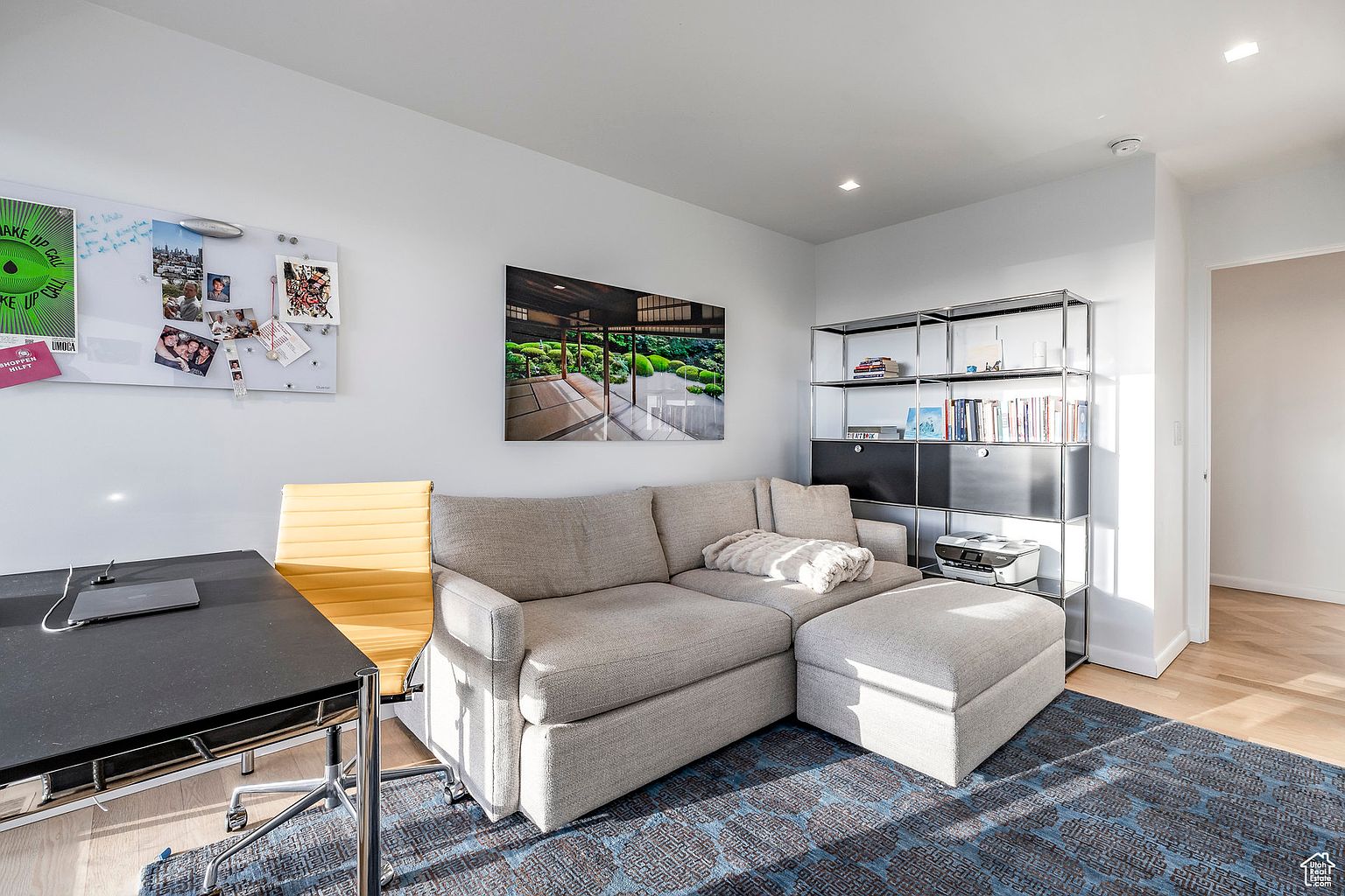 This interior shot showcases a well-lit office or study area featuring a comfortable gray sofa with an ottoman, a modern black desk with a yellow chair, and a metal shelving unit. A large piece of artwork hangs above the sofa, and a whiteboard is mounted on the wall, creating a functional and stylish workspace. The room is grounded by a patterned blue rug, adding a touch of color and texture.