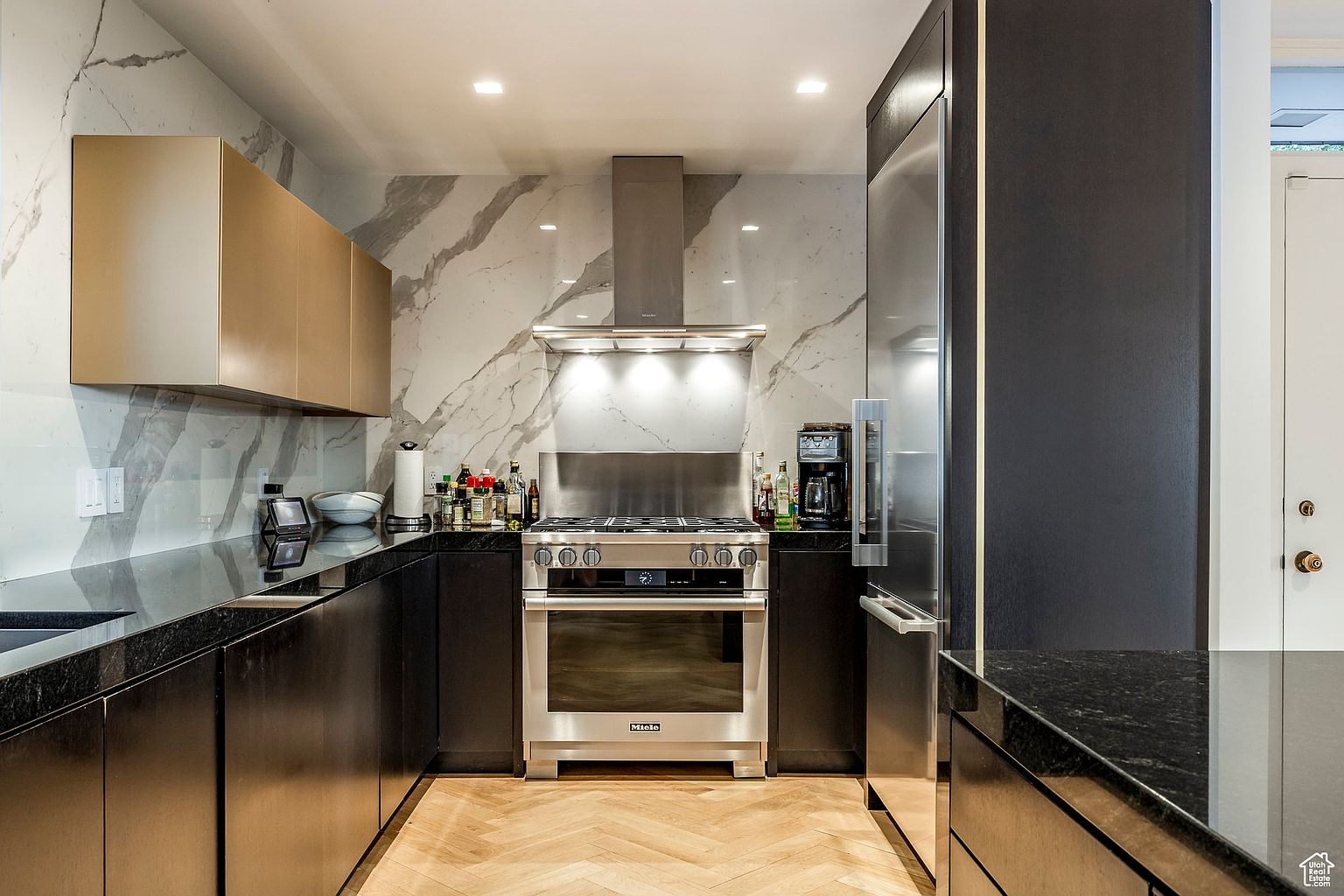 This is a modern kitchen with dark lower cabinets and gold upper cabinets. The countertops are black and the backsplash is a white marble with gray veining. Stainless steel appliances are featured, including a range with a hood and a refrigerator. The floor is a light wood in a herringbone pattern.