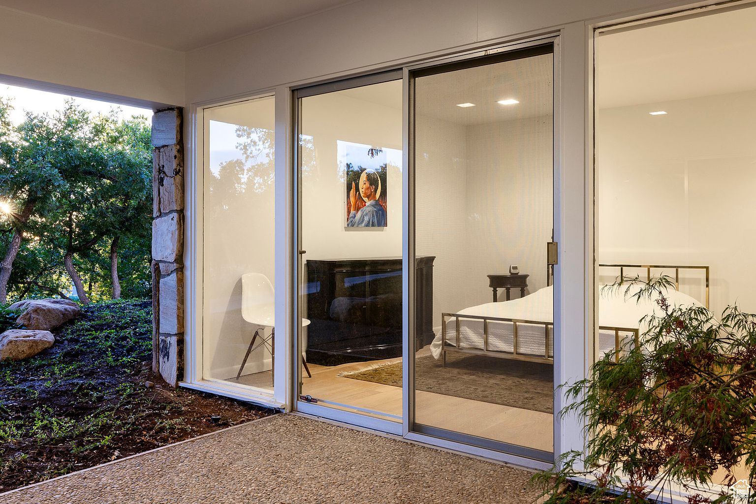 This image showcases a modern patio area seamlessly connecting the indoors with the outdoors. Large sliding glass doors provide access to a bedroom, while a stone-clad column adds a rustic touch. The patio features a textured concrete surface, and lush greenery surrounds the area, creating a tranquil and inviting atmosphere.
